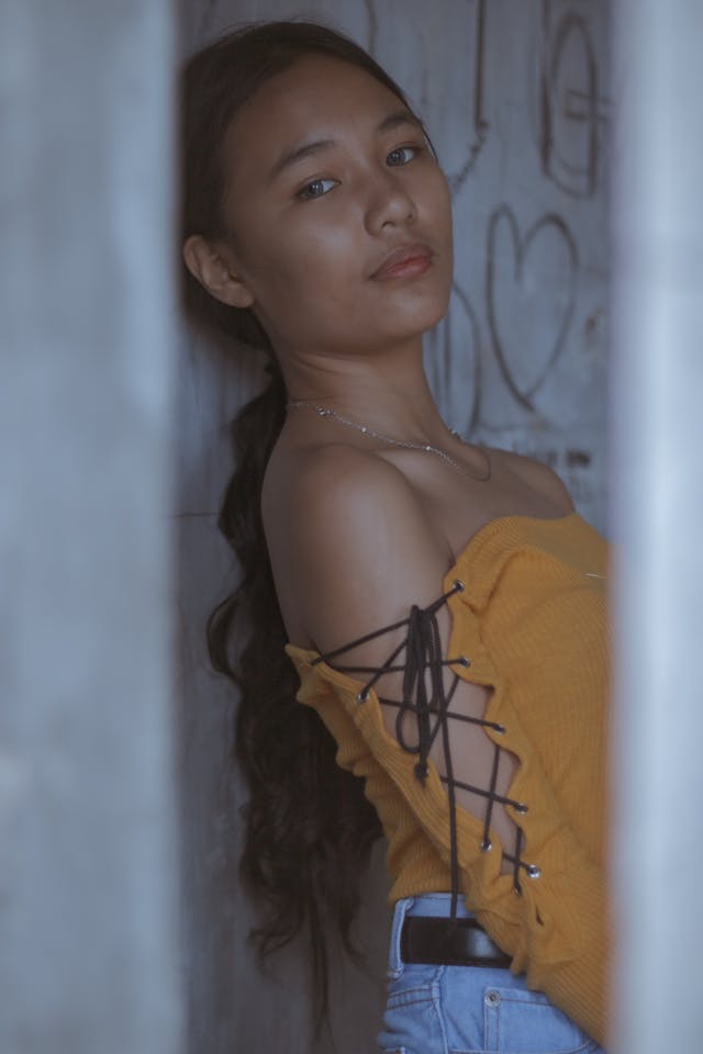 A woman wearing a yellow top leans against a wall.
