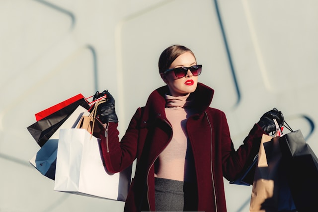 A woman in a dark red coat and sunglasses carries shopping bags with both hands. 
