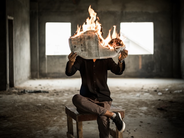 Uma pessoa se senta em um armazém abandonado e segura um jornal em chamas.