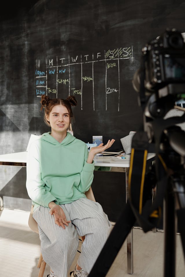 A girl in a green hoodie talks to a camera.
