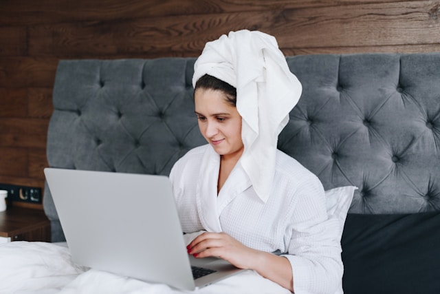 A woman wears a towel around her head and a robe and sits on the bed while browsing her laptop. 