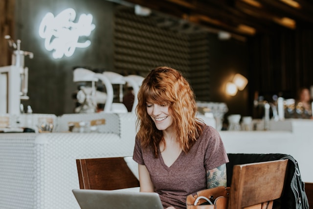 A woman in a coffee shop smiles as she types TikTok comments on her laptop. 
