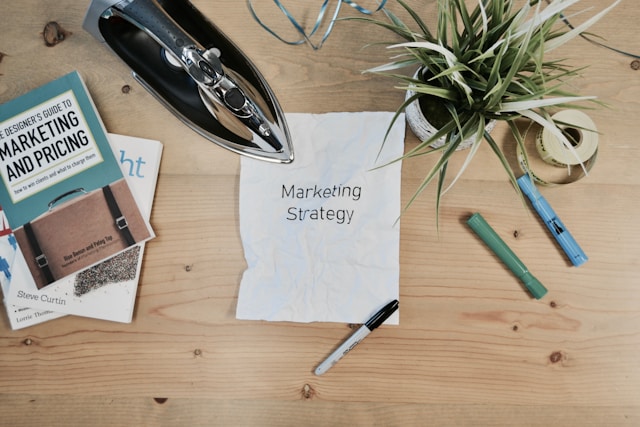 A table with an electric iron, a vase, pens, papers, and books about marketing strategies and pricing.
