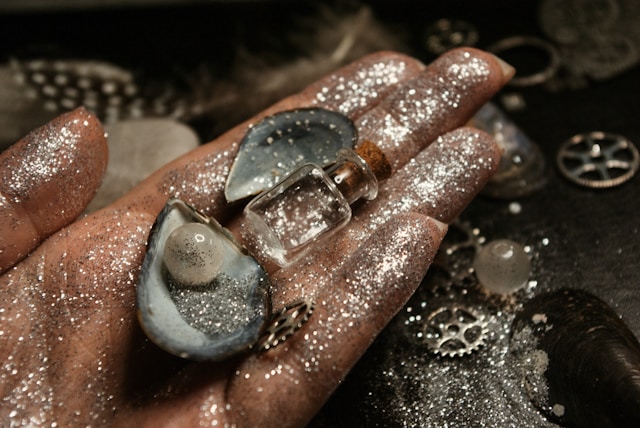 A hand covered in silver glitter holds a shell with a pearl in it. 
