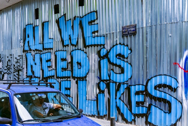 Blue graffiti on a corrugated sheet wall reads, “All we need is more likes.”
