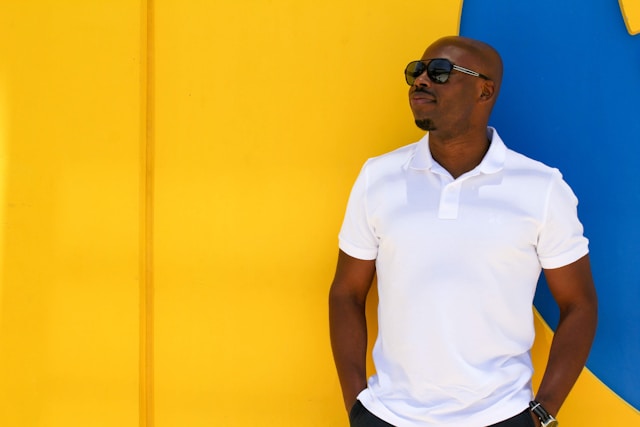 A man in a white shirt and dark sunglasses stands in front of a yellow and blue wall. 
