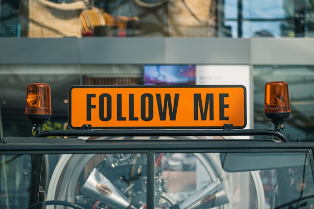 A black-and-orange sign that says, “Follow me,” is placed between two orange flashing lights. 
