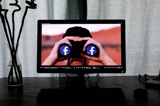 A computer screen displays a man looking through a pair of binoculars with the Facebook logo shown on both lenses. 
