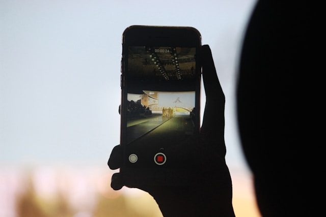 A person uses their phone to record a video of people walking down a runway.
