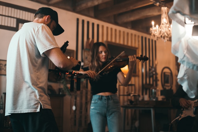 A man with a video camera records a woman playing the violin. 
