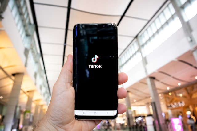 A person in public holds up their phone, which displays the TikTok logo.