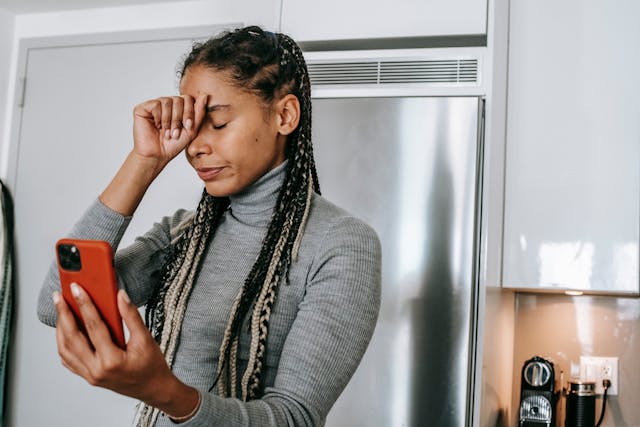 A woman holding a red phone puts one hand on her forehead.
