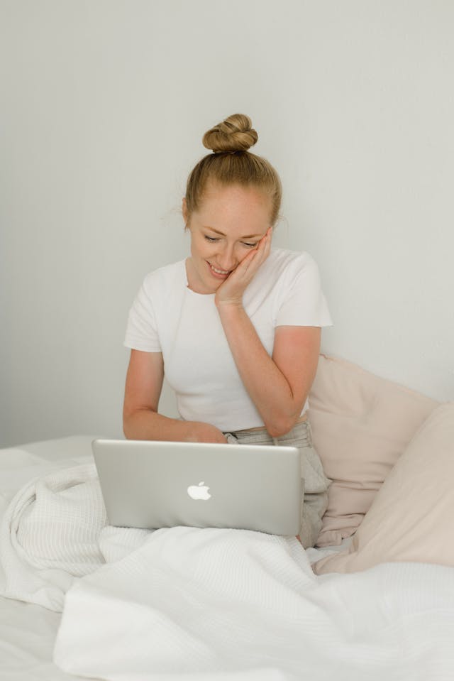 A woman sitting in bed smiles as she looks at her laptop.
