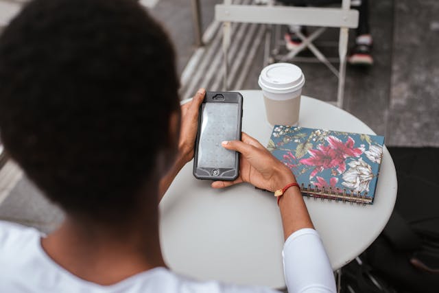 A woman uses her phone while sitting at a table with a phone and a cup of coffee.
