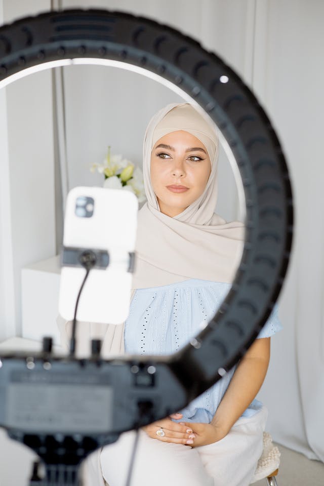 A phone in a ring light records a woman in a hijab.
