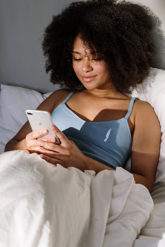 A woman smiles while using her phone in bed.
