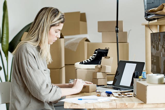 A woman surrounded by boxes types on her laptop.
