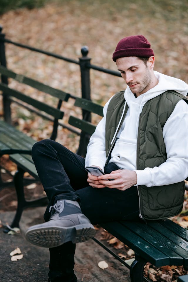 A man types on his phone while sitting on a park bench.
