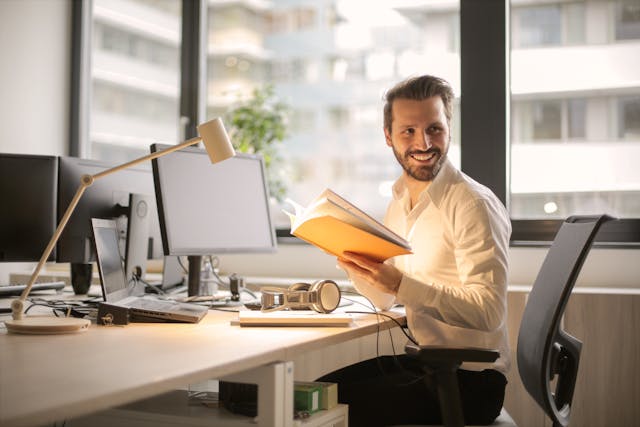 A man reads a book as he sits around a desk.
