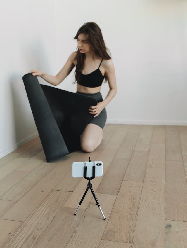 A phone on a tripod records a woman with a yoga mat.
