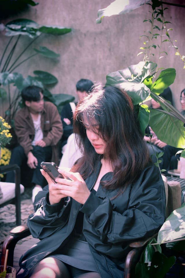 A woman sitting on a bench in a black jacket scrolls on her cell phone.
