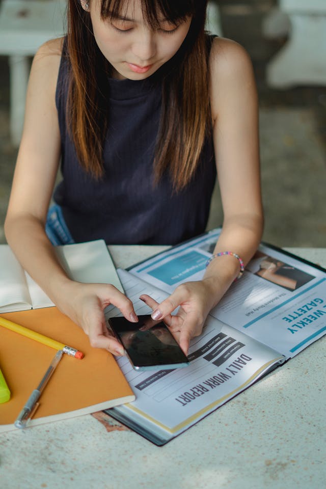 A woman rests her arms on books while she scrolls on her phone.