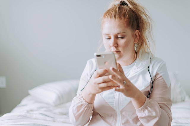 A woman with red hair sits on a white bed as she scrolls on her phone.
