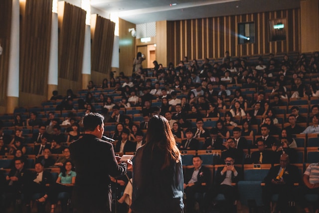 Two professional speakers stand in an auditorium in front of a big audience.