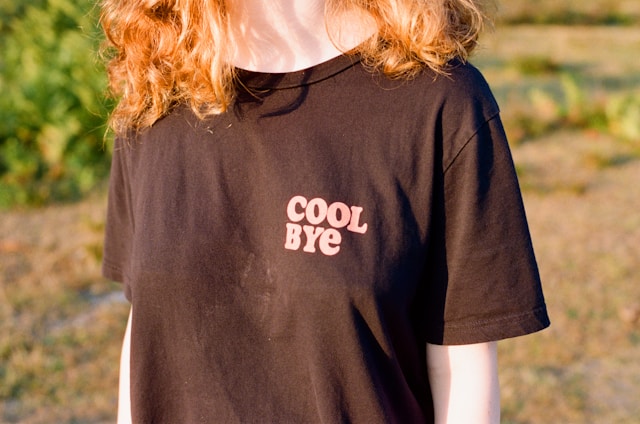 A woman with red hair wears a black shirt printed with the words, “Cool bye.”
