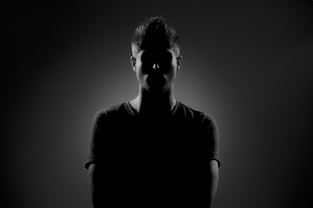 A man in a gray shirt stands in shadows to obscure his face. 
