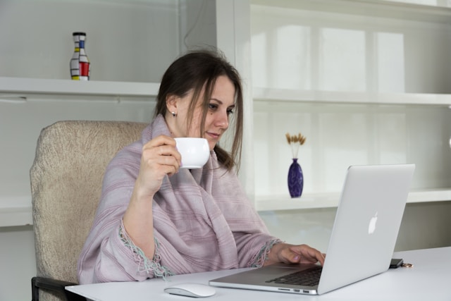 A woman wearing a blanket sits at a table and drinks from a cup while browsing videos on TikTok. 
