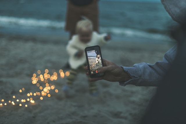 A father uses his phone to take a video of his child at the beach. 