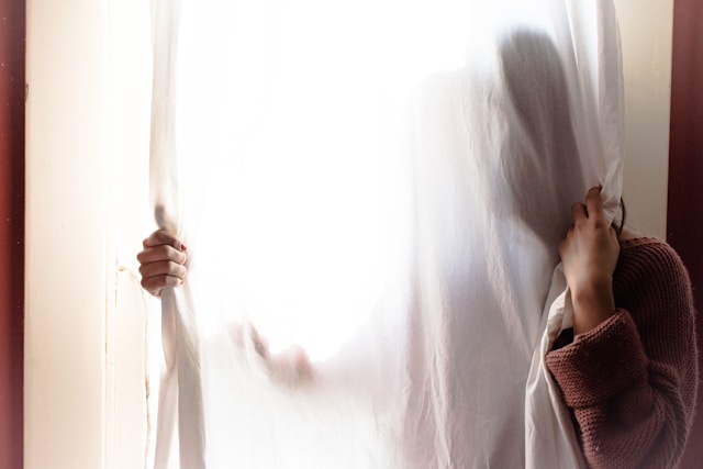 A woman in a peach sweater hides behind a sheer, white curtain. 
