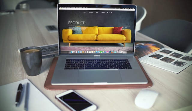 A laptop screen displays a living room product showcase featuring a yellow couch, throw pillows, and a pink rug. 