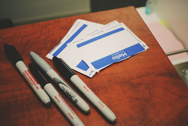 A stack of blank, blue, and white name tags are on top of a table beside some black markers. 
