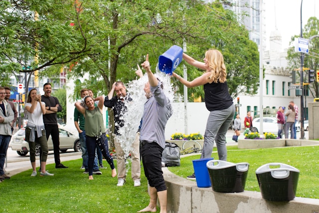 A woman pours a bucket of ice on a man while a small crowd cheers and takes videos with their phones.
