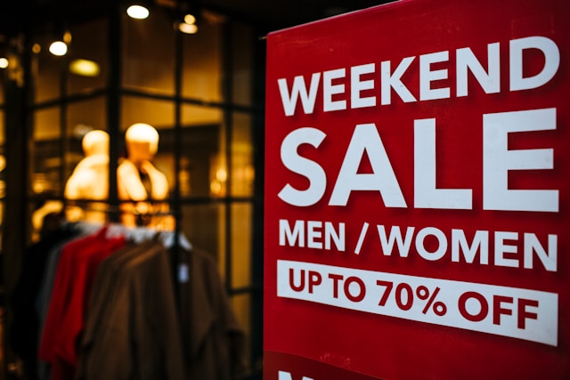 A store window displays a red and white sign that says, “Weekend sale, men/women up to 70% off.”
