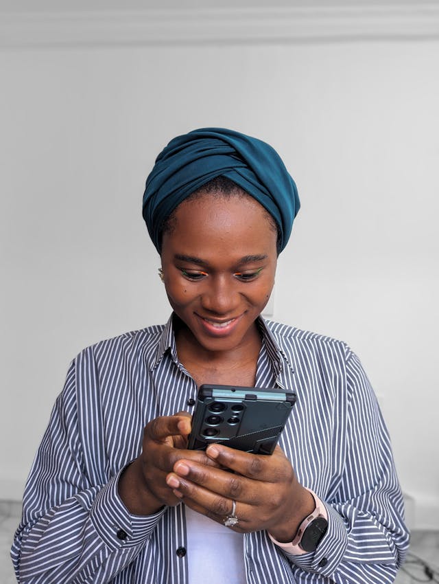 A woman in a head wrap smiles at her phone.
