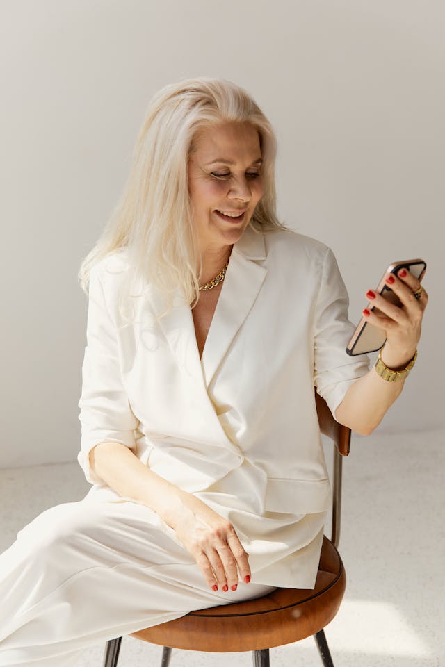 A woman with gray hair in a white suit smiles at her phone.
