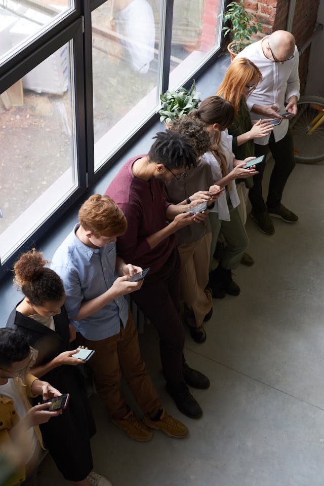 A variety of people line up against a wall and use their phones.
