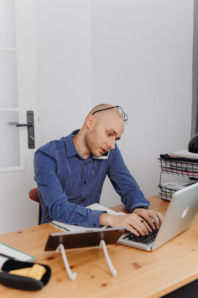 A man types on his laptop while he speaks on his cell phone.
