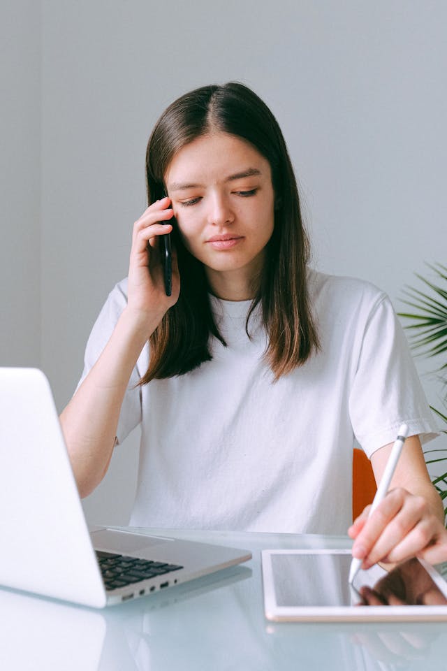 A woman uses her tablet with her phone at her ear.
