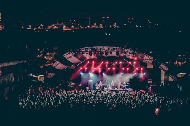 A big audience gathers around a stage at an outdoor concert. 
