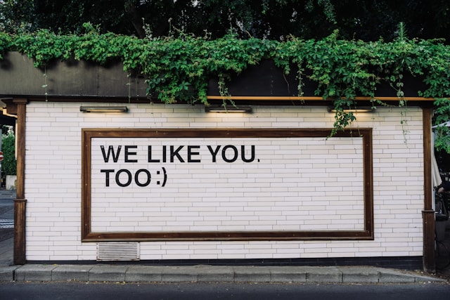 A sign on a white brick wall reads, “We like you, too.”
