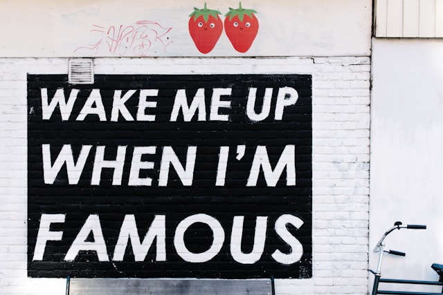 A spray-painted sign on a wall reads, “Wake me up when I’m famous.”
