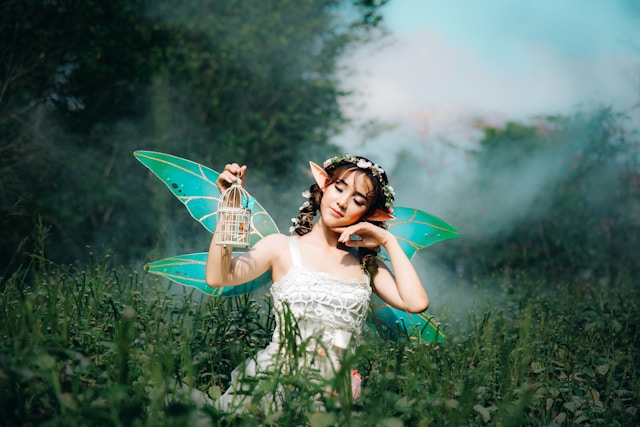 A woman in a fairy costume carries a tiny cage in a field of wildflowers.