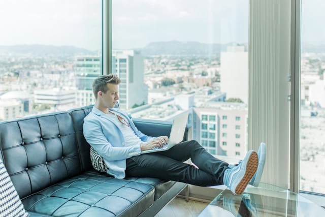 A young professional sits on a couch while checking on his business on TikTok using his laptop.
