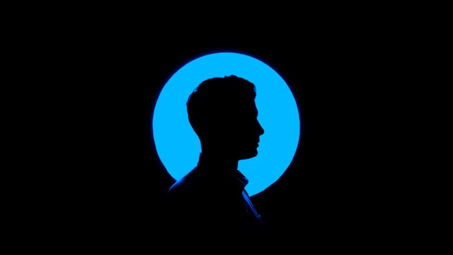 A dark silhouette of a man standing in front of a blue circle of light. 
