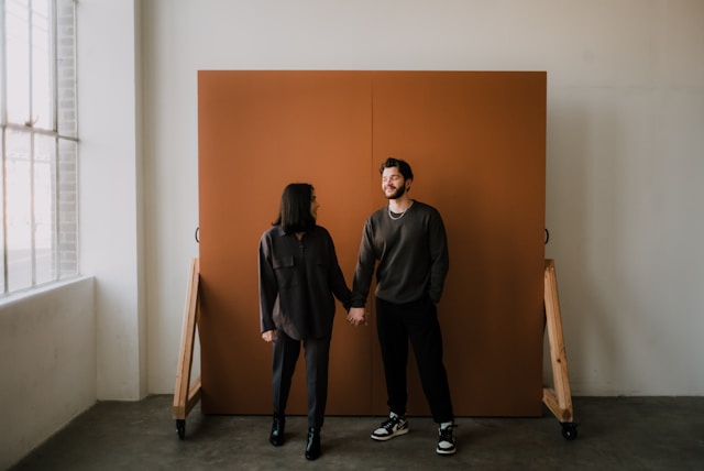 A couple stands in front of a solid bronze backdrop for a photoshoot. 
