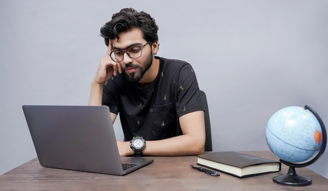 A man touches his temple with a finger while he browses on his laptop. 
